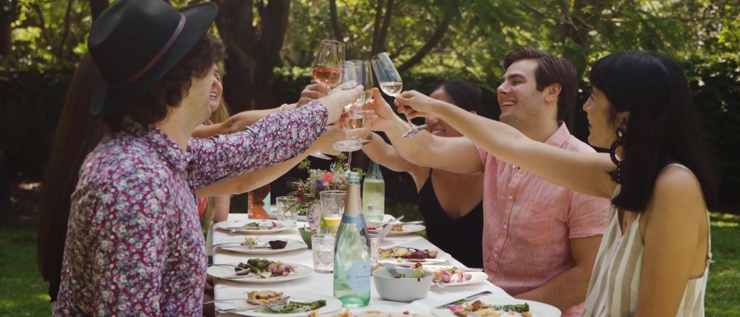 Friends celebrate a toast while having a meal seated at a table in a garden.